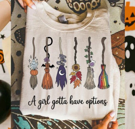 T-shirt with Halloween-themed broom designs and text 'A girl gotta have options' held against a colorful Halloween-themed background.