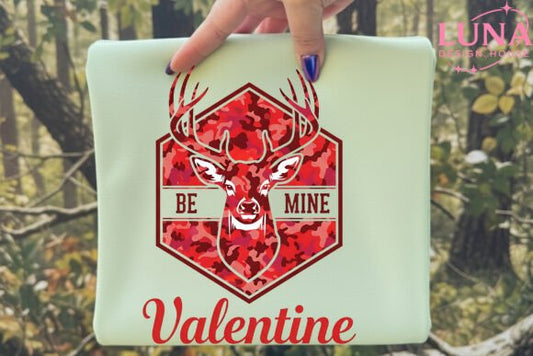 Laptop with a red deer design and 'Be Mine Valentine' text held by a hand in a forest setting.