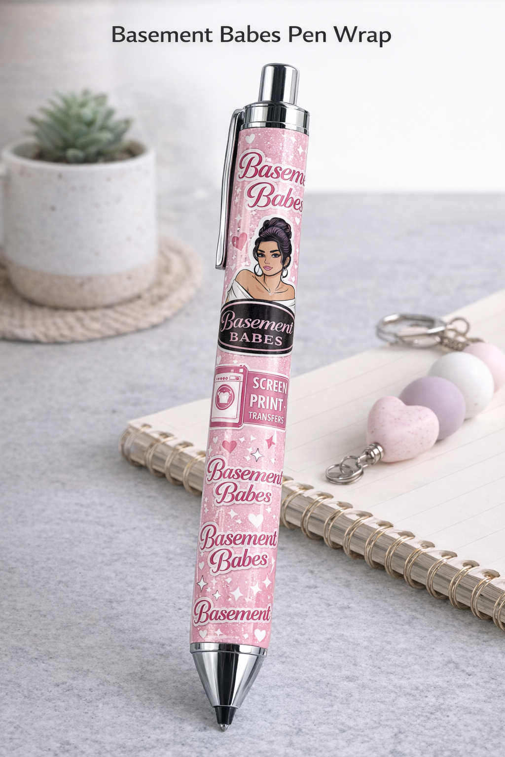 Pink pen with 'Basement Babes' branding on a gray surface with a plant and notebook in the background.